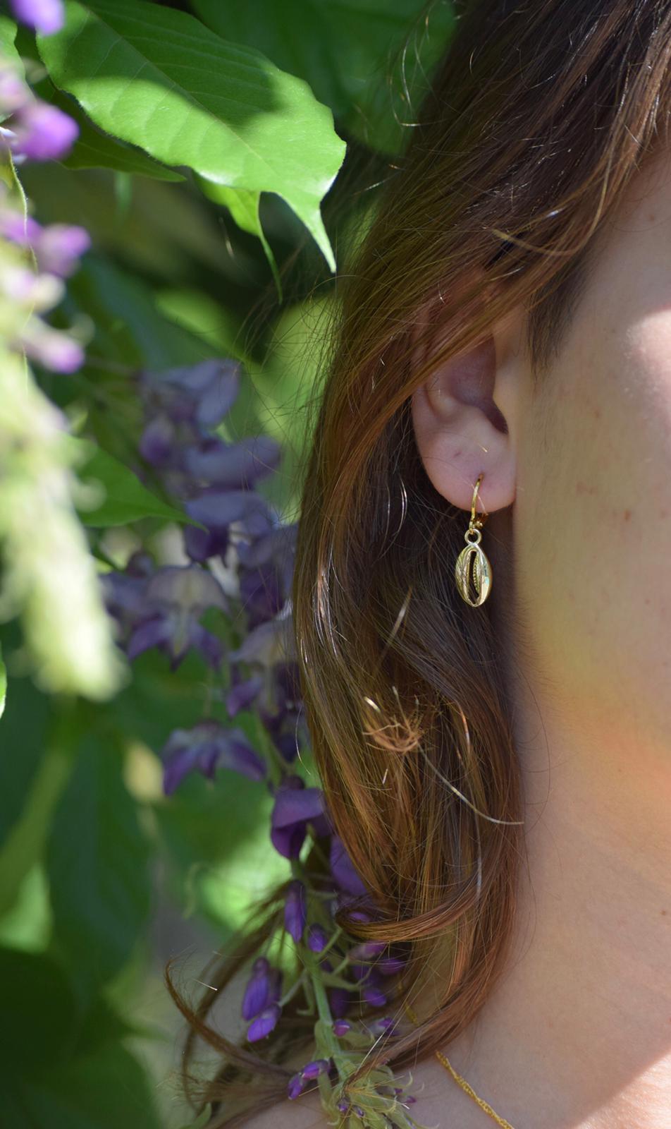 model staat in de natuur en draagt goudkleurige schelp oorbellen, het is een zomerse foto.